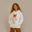 A young girl with long curly light brown hair wearing a cream-colored fleece hoodie with a 3D cat plush toy attached to the chest, paired with brown pants, standing against a neutral textured wall with dappled sunlight.