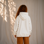 A person viewed from behind wearing an oversized off-white hooded sweatshirt and brown pants, standing against a light neutral backdrop with dappled sunlight and shadows.