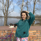 Woman in teal hoodie with a dog on a leash by a brick wall and trees.
