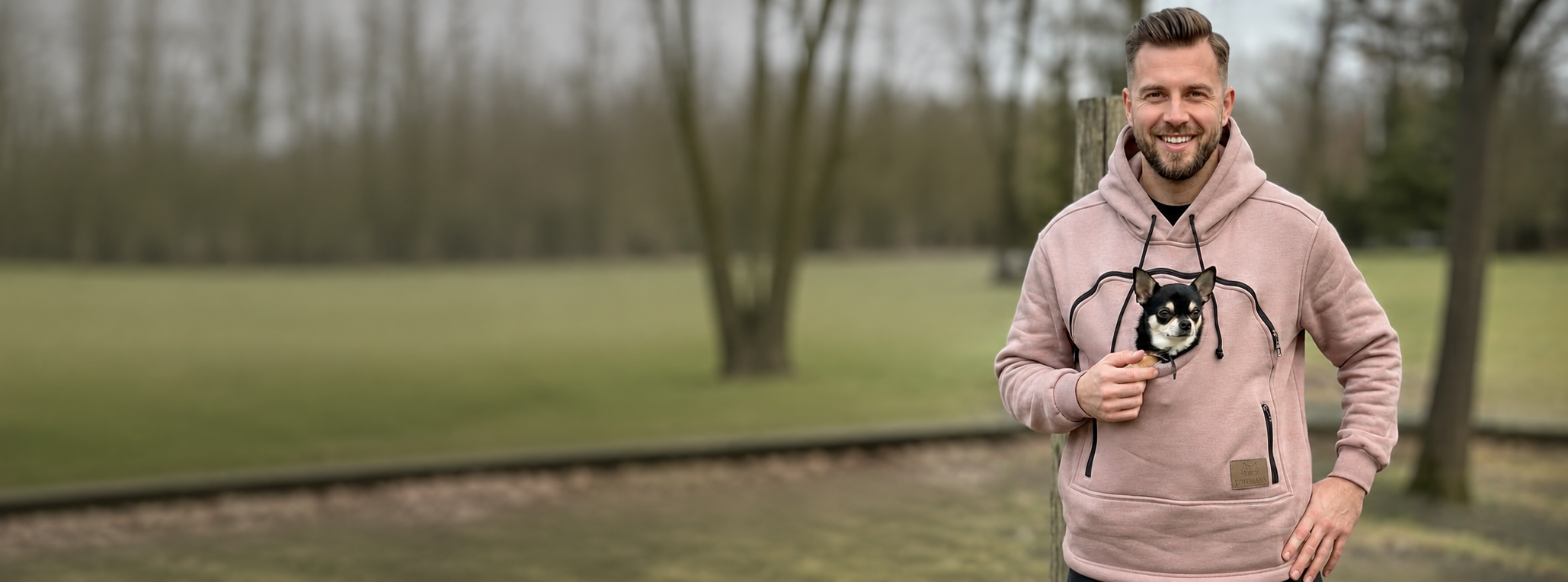 A man wearing a beige dog hoodie with a black dog, stands outdoors in a park.