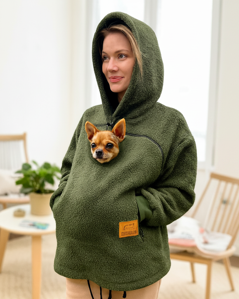 A woman wearing an olive green sherpa hoodie with a built-in pocket holding a small dog, standing in a bright, modern room with wooden chairs and a small table.
