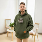 Man wearing a green khaki hoodie with a cat face design in a room with light-colored walls and wooden furniture.