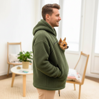 Man wearing a green Khaki fleece hoodie with a dog peeking out of the pocket in a room with light-colored walls and wooden furniture.