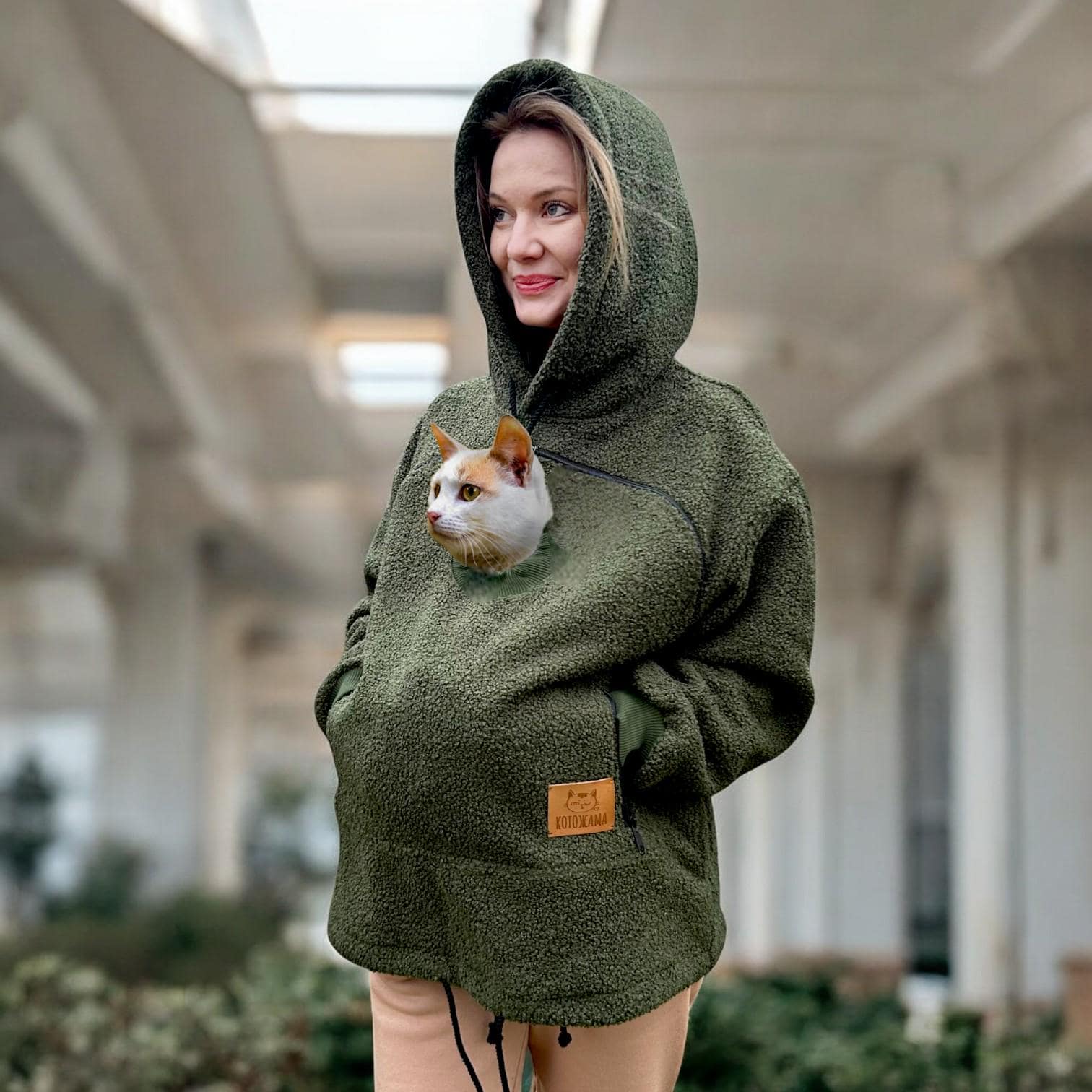 Person wearing a Khaki Sherpa hoodie with a pocket containing a cat, standing indoors.