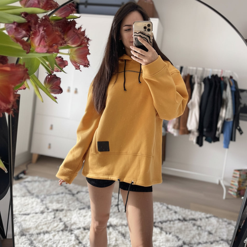 Person wearing a Mustard hoodie taking a mirror selfie in a room with flowers and clothing.