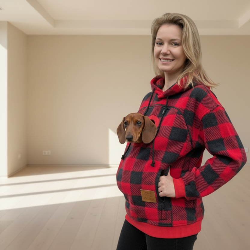 Woman wearing a red and black checkered hoodie with a dog peeking out from the front pocket in an empty room.