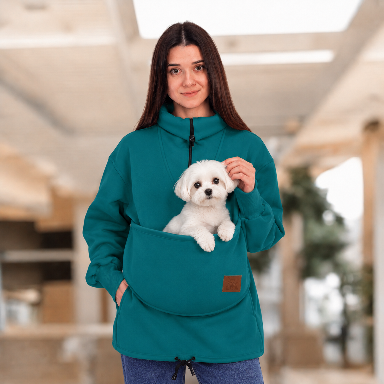 Women wearing a teal sweatshirt with a white maltipoo dog in pouch, standing indoors.