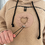 Close-up of a beige fleece hoodie with a drawstring and heart-shaped pocket patch.