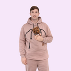 Person wearing a beige hoodie with a built-in dog carrier holding a small dog on a white background