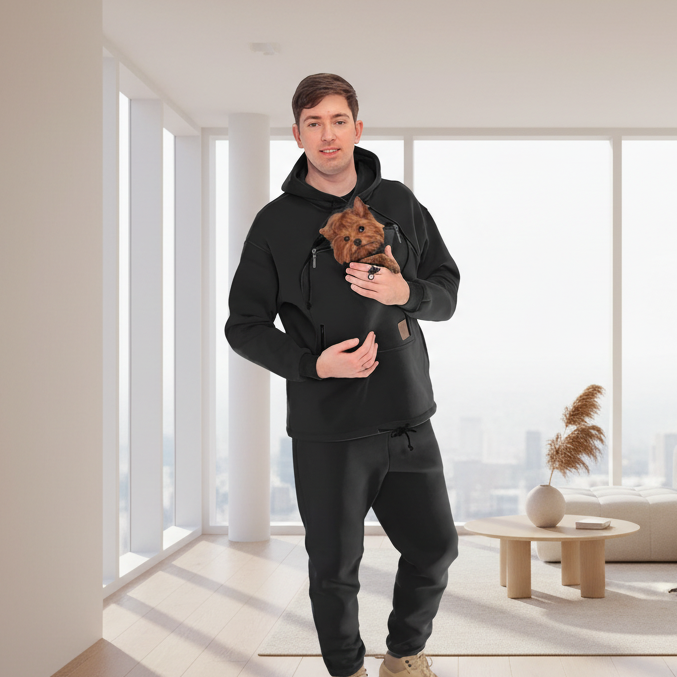 Man holding a small dog in a black hoodie with a pocket, standing in a modern living room. Основне зображення