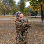 Person wearing a camouflage hoodie holding a gray cat outdoors with trees and fallen leaves in the background.
