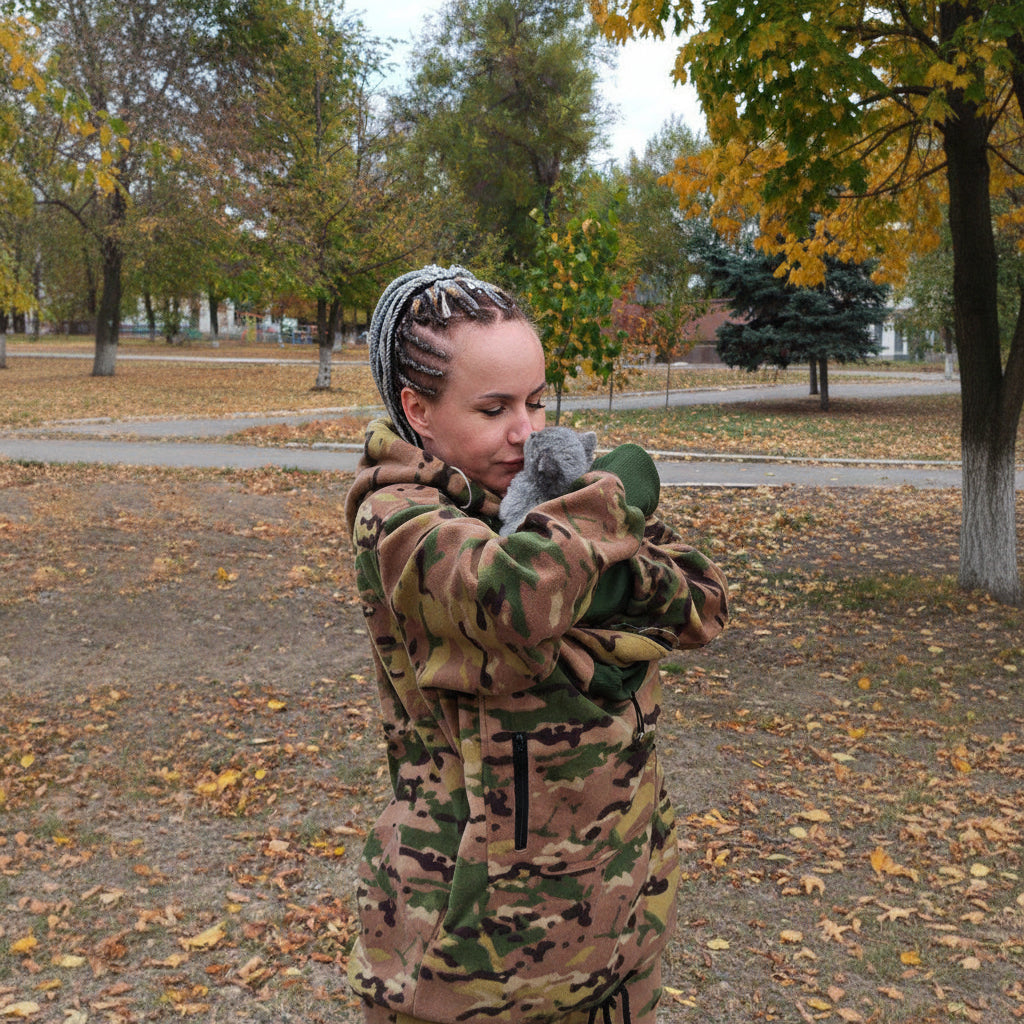 Person wearing a camouflage hoodie holding a gray cat outdoors with trees and fallen leaves in the background.