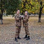 Two people in camouflage outfits standing outdoors with trees in the background.