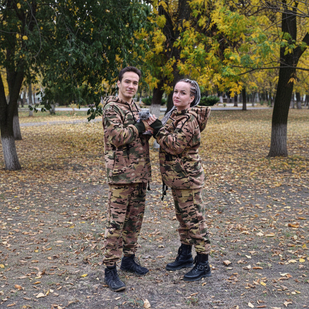 Two people in camouflage outfits standing outdoors with trees in the background.