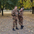 Two people in camouflage outfits standing outdoors with trees and fallen leaves in the background.