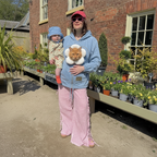 A woman wearing a light blue hoodie with a cat wearing a white flower collar and neck warmer, paired with pink gingham side-slit pants, stands outdoors in front of a brick building holding a toddler.
