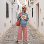 A woman stands in a narrow white-walled cobblestone alley wearing a light blue hoodie with a cat-shaped patch, red-and-white gingham cropped pants, red slip-on shoes, and a beige hat. She carries a red patterned tote bag.