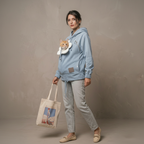 A woman modeling a light blue hoodie with a built-in pet carrier pocket containing an orange cat, paired with light gray joggers and beige flats, while holding a canvas tote bag against a textured beige studio backdrop.