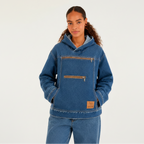 A woman wearing a blue hooded denim jacket with brown zipper details and a tan leather patch on the lower front, paired with matching blue jeans, against a plain white studio background.