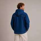 A man wearing a blue hooded denim jacket, shown from the back and side against a neutral gray studio background.