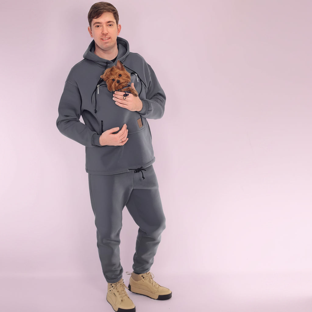 Man wearing a gray hoodie and pants holding a small dog, standing against a white background