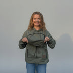 A woman wearing an olive green hooded jacket with a drawstring pouch on the chest, holding it open to show the interior. She is wearing blue jeans and standing in front of a beige fabric backdrop.
