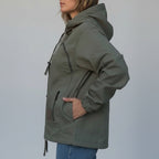 A person wearing an olive green hooded jacket with multiple zippered pockets, shown from the side against a textured beige backdrop.