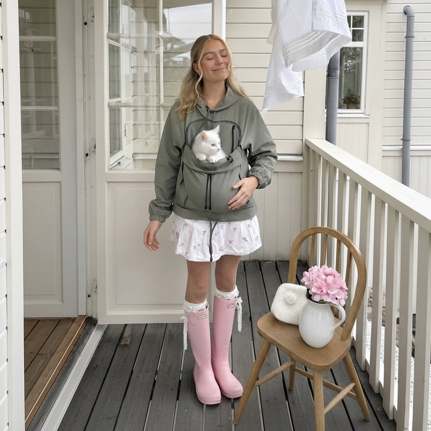 A woman stands on a porch wearing an olive green hooded sweatshirt with a front pet carrier pocket containing a white cat. She is also wearing a white ruffled skirt, pink rain boots, and holding a small white purse.