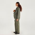 A woman in profile wearing an olive green utility jacket and matching wide-leg trousers, holding a small brown dog against her chest. She is standing against a plain white studio background.