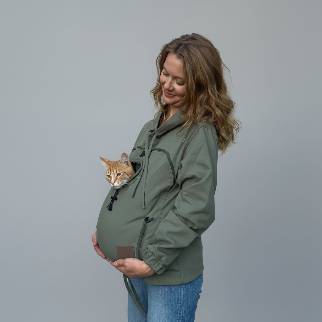 A pregnant woman wearing an olive green cat carrier jacket with an orange tabby cat peeking out from the front pouch, standing against a beige wrinkled backdrop.