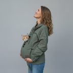 A pregnant woman wearing an olive green maternity carrier jacket with a cat peeking out of the front pouch, set against a solid gray studio background.