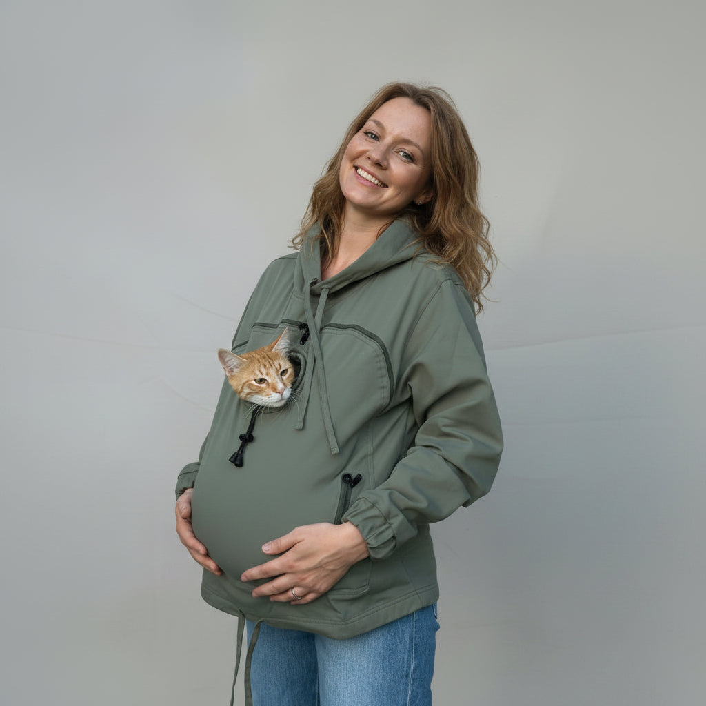 A pregnant woman wearing an olive green hoodie with a cat peeking out of the chest pocket, standing against a wrinkled beige backdrop. Main image