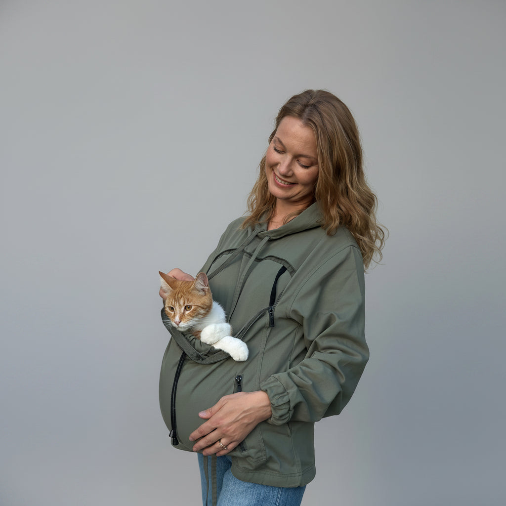 A woman wearing an olive green hooded jacket with a cat carrier pocket, holding an orange and white cat against her stomach, set against a wrinkled beige backdrop.