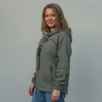 A woman wearing an olive green hooded pullover jacket with drawstrings and elastic cuffs, paired with blue jeans, standing in front of a wrinkled beige backdrop.