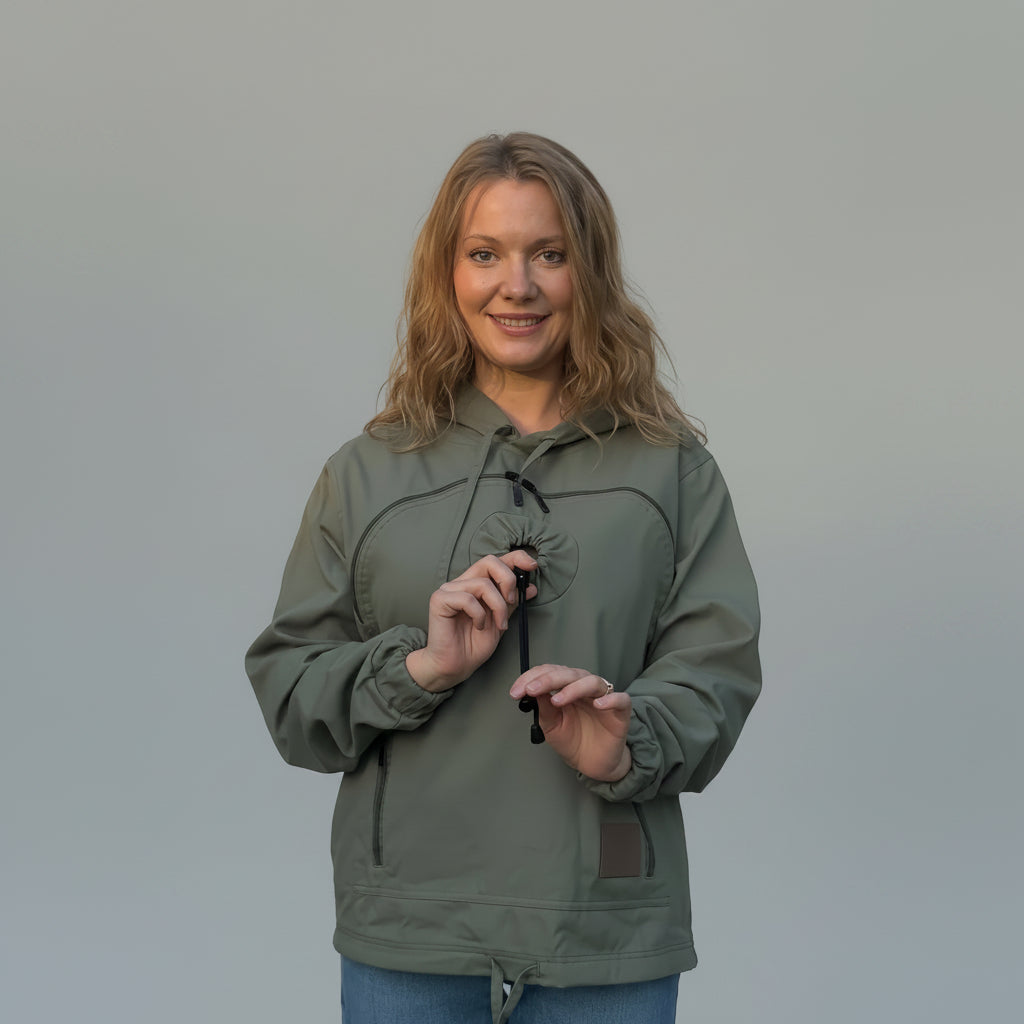 A woman wearing an olive green hooded pullover jacket with drawstrings, paired with blue jeans, standing against a textured beige backdrop.