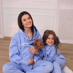 Woman and child in matching light blue hoodies with a small dog, sitting on a light-colored floor.