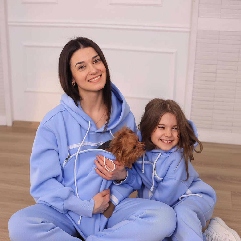 Woman and child in matching light blue hoodies with a small dog, sitting on a light-colored floor.