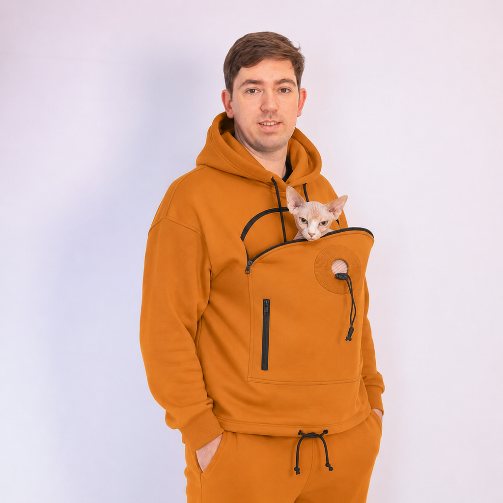 Man wearing an orange hoodie with a built-in pet carrier, holding a small white cat.