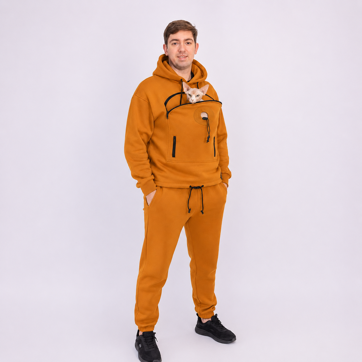 Man wearing a mustard yellow tracksuit with a small cat peeking out of the front pocket on a white background Main image