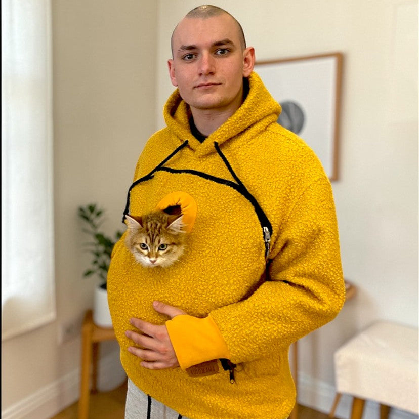 Person wearing a orange boucle hoodie with a cat peeking out of the pocket, standing in a room.