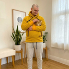 Man in orange fleece hoodie and light gray sweatpants holding a cat indoors.