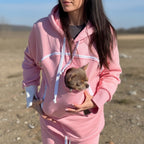 Person wearing a pink set with a small dog peeking out of the front pocket, standing on a beach.