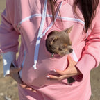 Person wearing a pink hoodie with a small dog peeking out of the front pocket, standing on a beach.