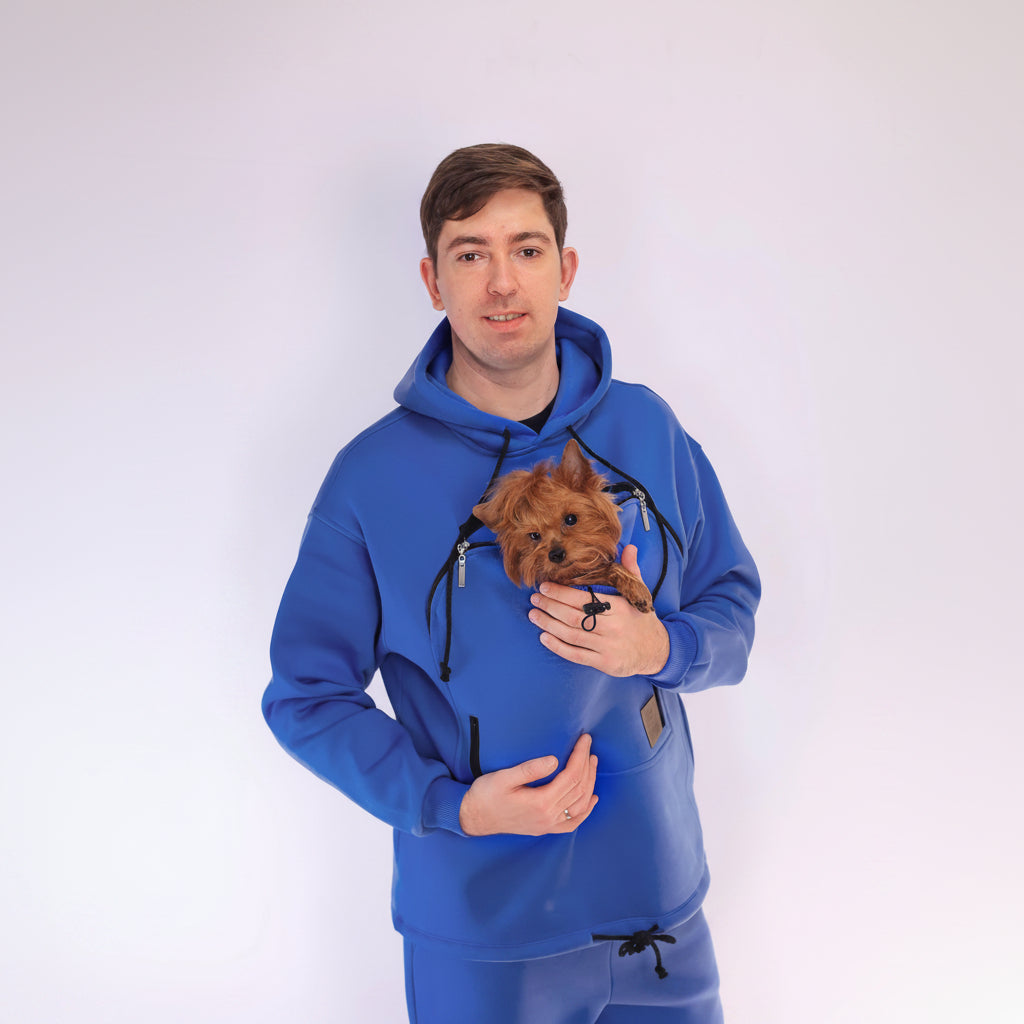 Man wearing a royal blue hoodie holding a small dog against a white background