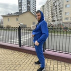 Man in blue tracksuit holding a cat in a carrier bag on a sidewalk with buildings in the background