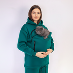 Woman wearing a teal hoodie with a cat carrier holding a gray cat on a white background