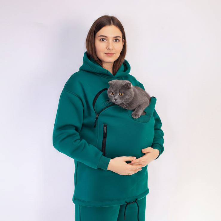 Woman wearing a teal hoodie with a cat carrier holding a gray cat on a white background