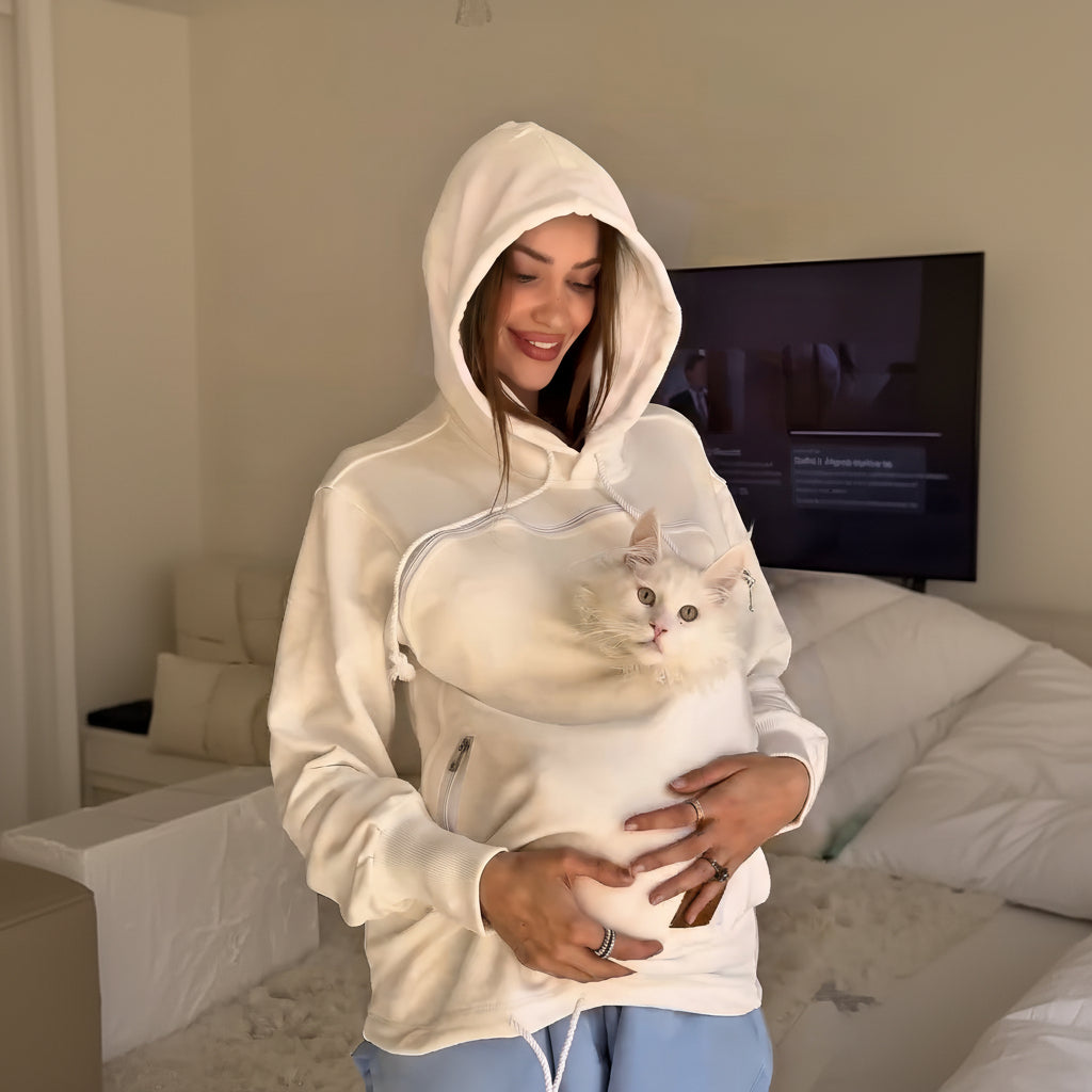 Person wearing a white hoodie with a cat inside, standing in a neutral indoor setting.