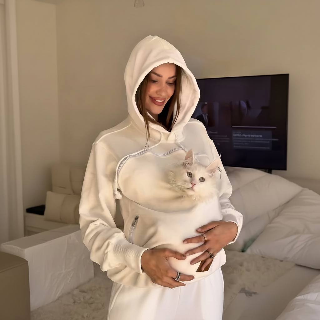 Woman wearing a white hoodie with a pouch, holding a cat inside the pouch, in a bedroom setting.