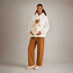 A woman stands against a plain light gray studio background wearing a white hoodie with a cat stuffed inside the front pocket, paired with wide-leg brown pants and cream slip-on shoes.
